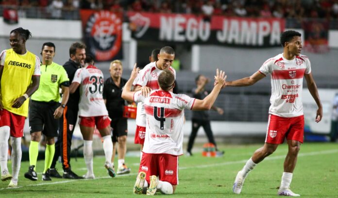 Dois jogadores de futebol comemorando um gol com um abraço em campo durante uma partida do Brasileirão enquanto outros passam ao fundo.