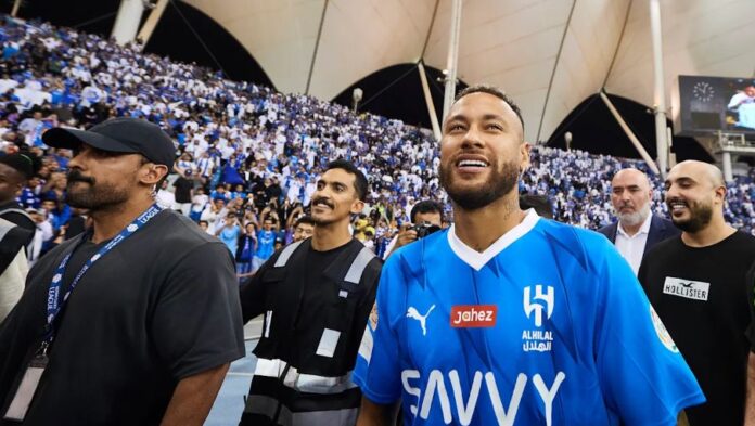 Um homem com uma camisa de futebol azul do Al-Hilal com o número 14 se destaca na multidão de fãs de esportes.