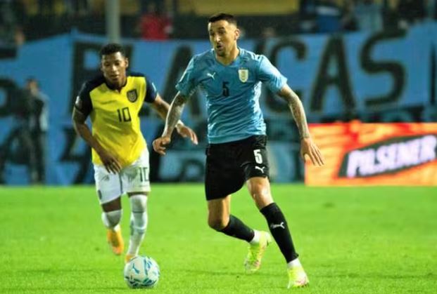 Dois jogadores de futebol no meio de uma partida, sendo um deles com a camisa do Equador perseguindo a bola controlada por um jogador de camisa azul claro.